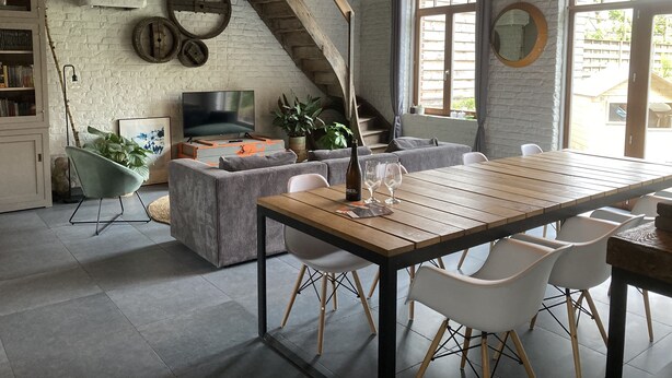 Un salon chaleureux avec une table en bois, des chaises blanches, un téléviseur et un escalier.