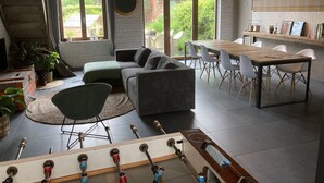 Un salon confortable avec un canapé, une table à manger et des chaises, ainsi qu’une table de baby-foot. De grandes fenêtres offrent une vue sur la verdure environnante.