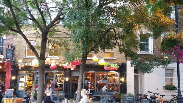 An outdoor cafe with tables and chairs, bicycles parked nearby, and a building with large windows in the background. - Fabulous one double bedroom flat in garden setting by the river in Battersea (London)