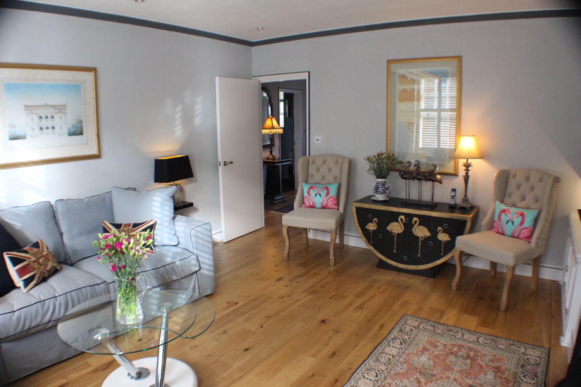 En stue med en grå sofa, et glasbord, en beige lænestol, et sort skænk med flamingo dekorationer, og en tæppe med et mønster.