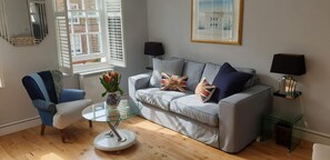A living room with a grey sofa, a blue armchair, a glass coffee table, and a vase with flowers. There are two black lamps on side tables, and a framed picture on the wall.
