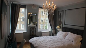 A bedroom with a large bed, a chandelier, a mirror, and two windows with curtains. - Fabulous one double bedroom flat in garden setting by the river in Battersea (London)