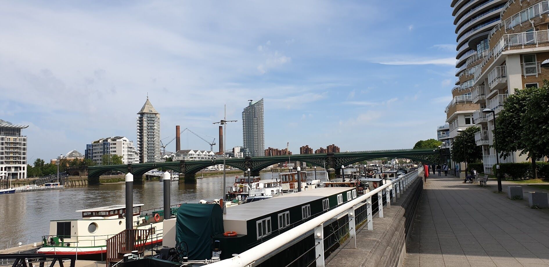 A riverside walkway with a bridge, boats, and buildings in the background.