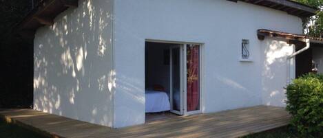 A single-story building with a white exterior, a wooden deck, and a sliding glass door leading to a room with a bed.