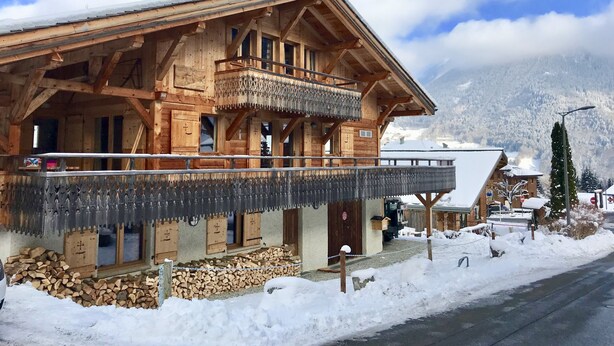 A wooden chalet with a balcony, wooden logs stacked outside, and a snowy mountain backdrop. - Superb chalet: 50m to ski lift. 6 large bedrooms, sleeps 12, sauna & steam rooms (Saint-Jean-D'aulps)