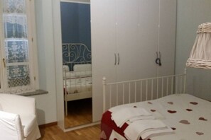 A bedroom with a bed, a wardrobe, a window with lace curtains, and a bedside table.