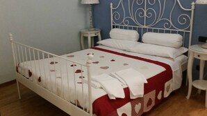 A white metal bed with a red and white patterned bedspread, two white pillows, a white bedside table with a lamp, and a small round table with a lamp.