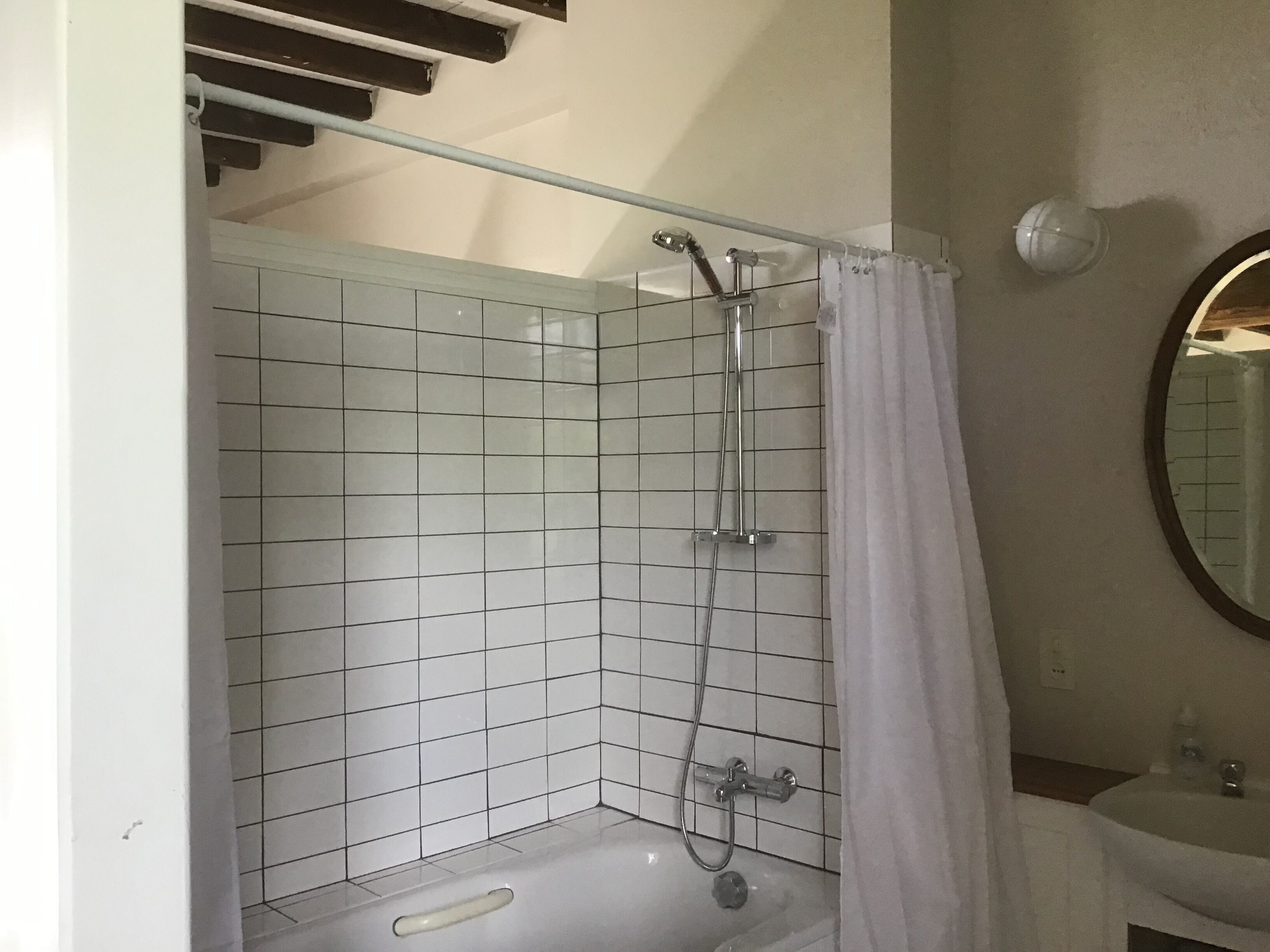 Ducha con puerta de cristal, pared con azulejos blancos y un lavamanos pequeño con espejo.