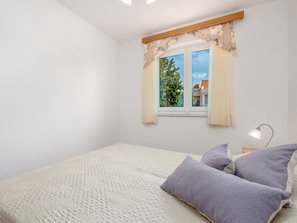 Un dormitorio con cama, almohadas, mesita de noche, lámpara y ventana con cortinas.