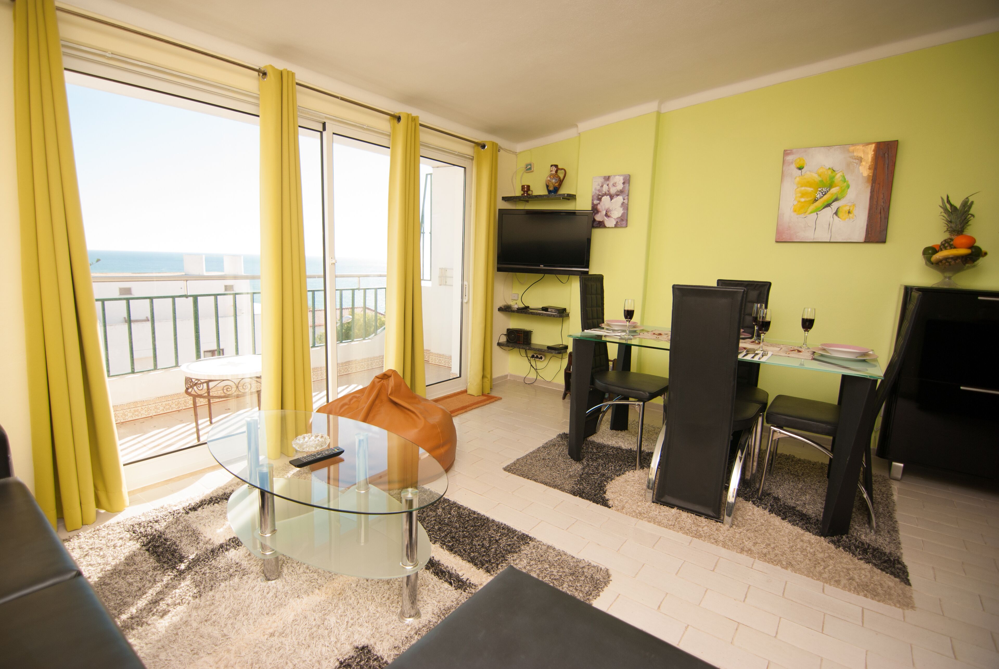 A modern living room with a glass coffee table, a sofa, and a dining area with a glass table and chairs. There is a TV mounted on the wall and a view of the sea through large windows.