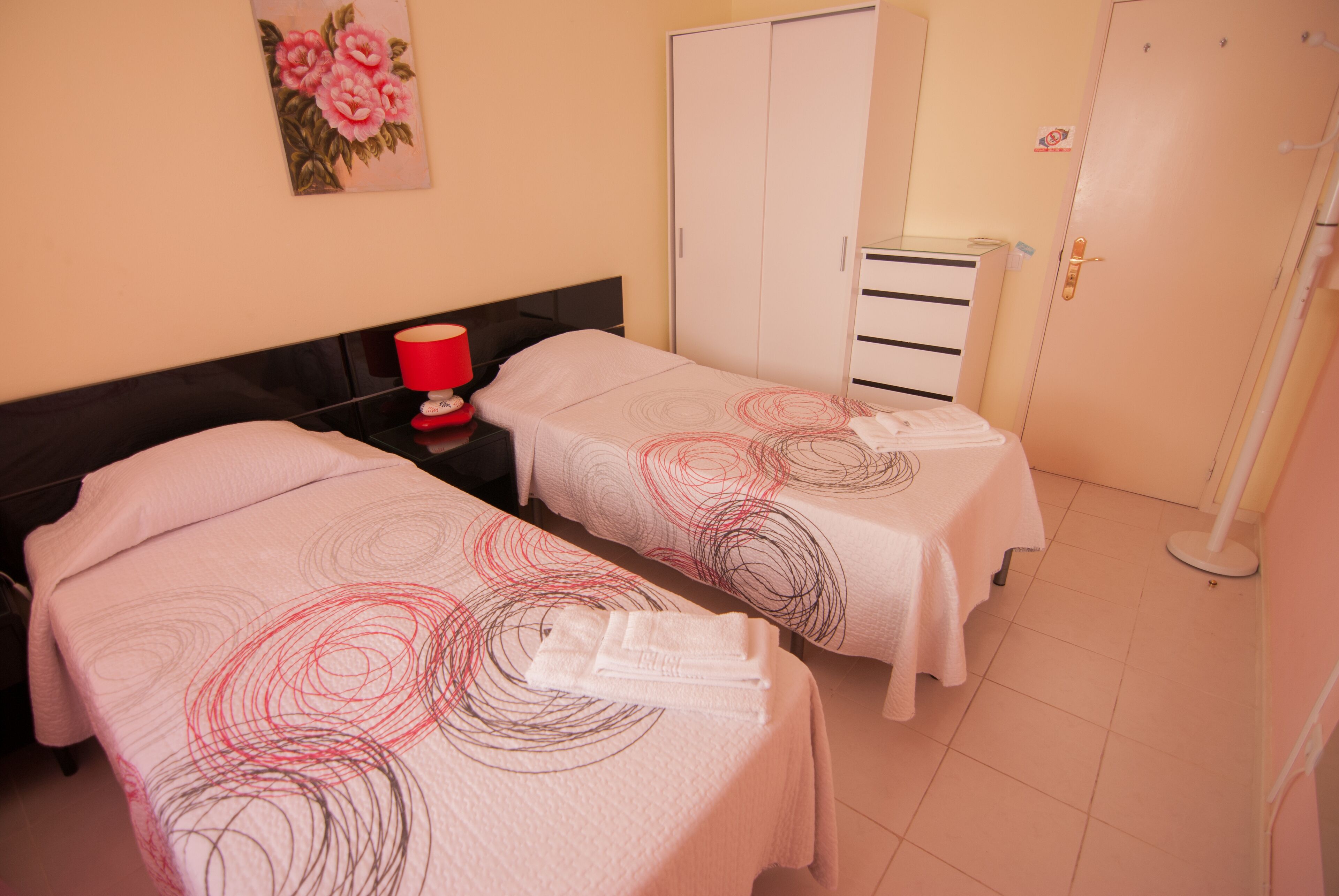 A hotel room with two beds, a wardrobe, a bedside table with a lamp, and a painting on the wall.