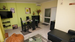 Un salon avec une table basse en verre, un fauteuil poire marron, un canapé noir, un coin repas avec une table et des chaises, un téléviseur fixé au mur et un vase avec des fleurs sur une table d’appoint.