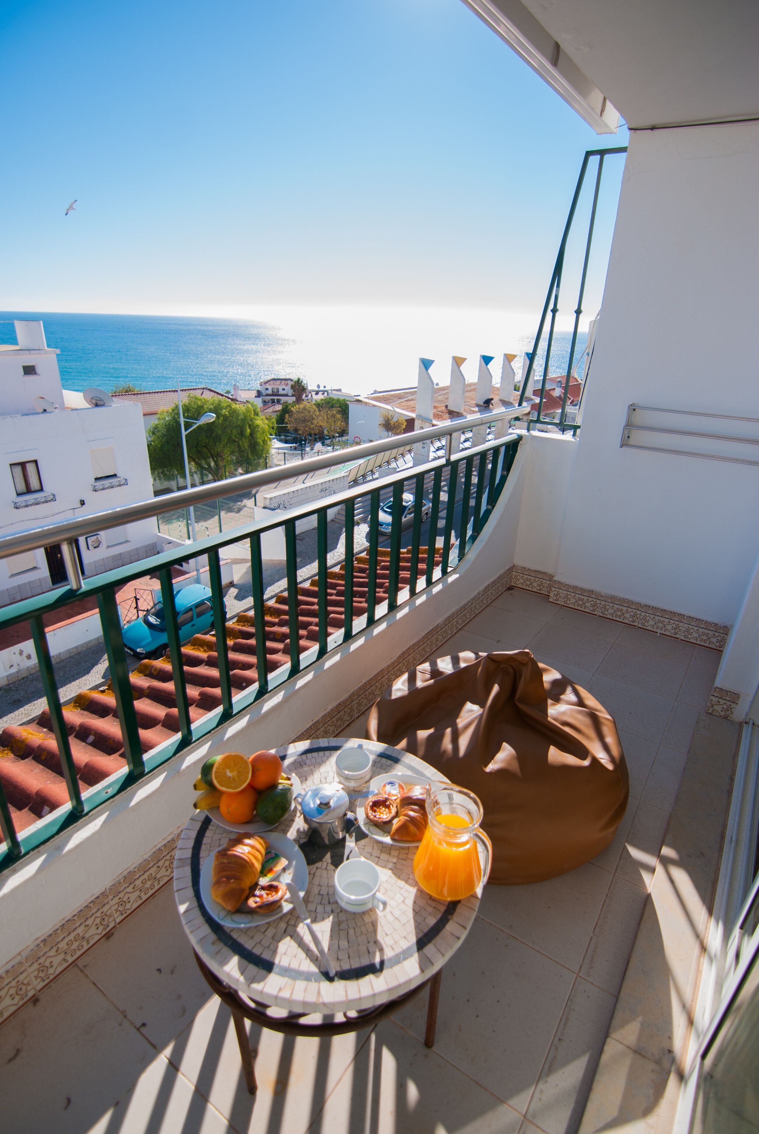 A balcony with a table set for breakfast, a bean bag, and a view of the sea and buildings.