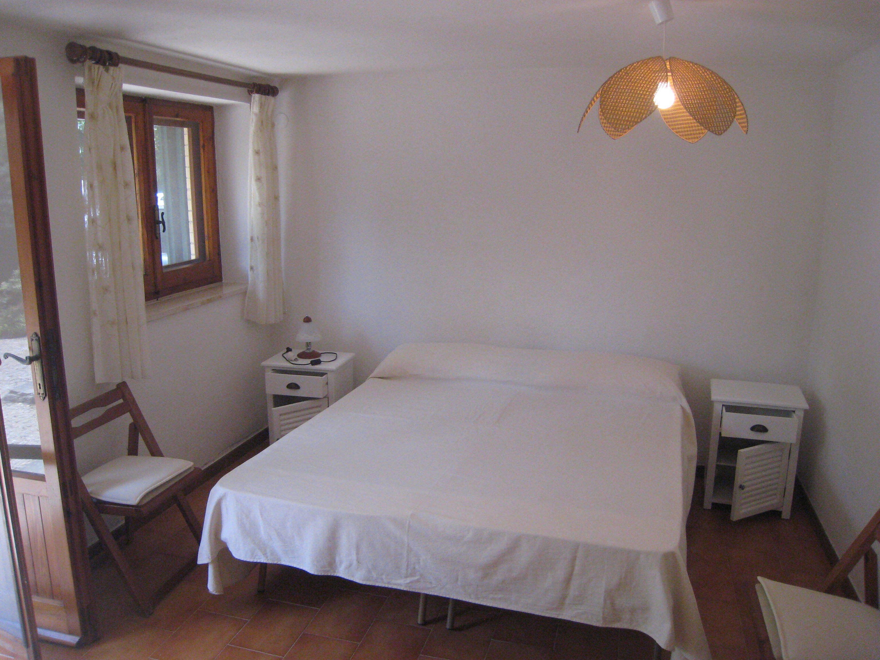 A bedroom with a bed, a nightstand, a chair, a window with curtains, and a hanging light fixture.