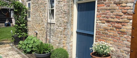 Un bâtiment en briques avec une grande fenêtre et une porte. On y voit des plantes en pot et une petite table sur laquelle est posé un objet décoratif.