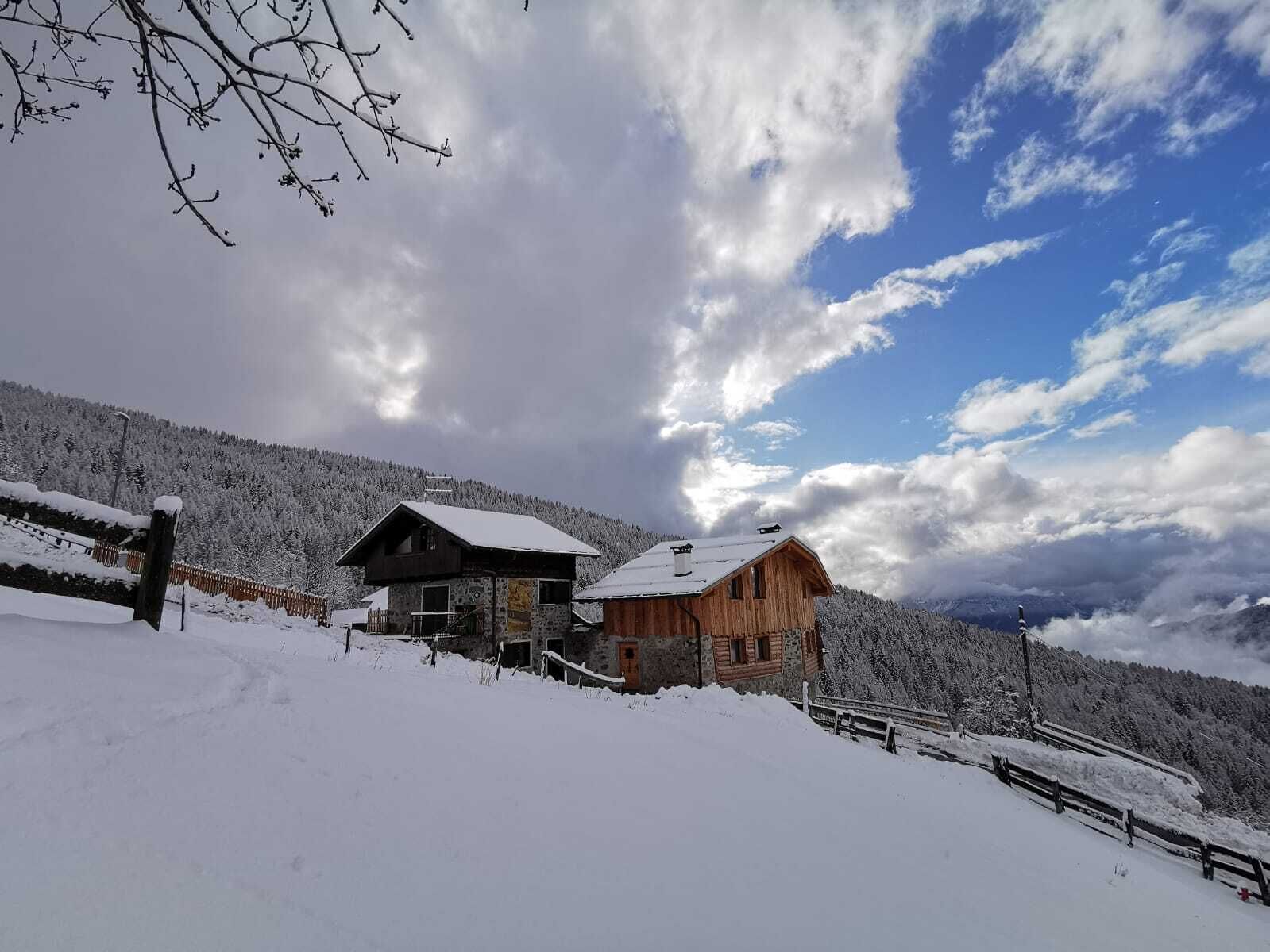 Une cabane en bois, dont le toit est recouvert de neige, entourée d’un paysage enneigé et adossée à une montagne boisée.