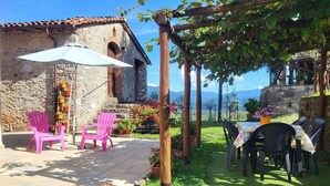 Un edificio de piedra con tejado de baldosas, mesas y sillas de plástico en la terraza, y una pérgola cubierta de plantas trepadoras.