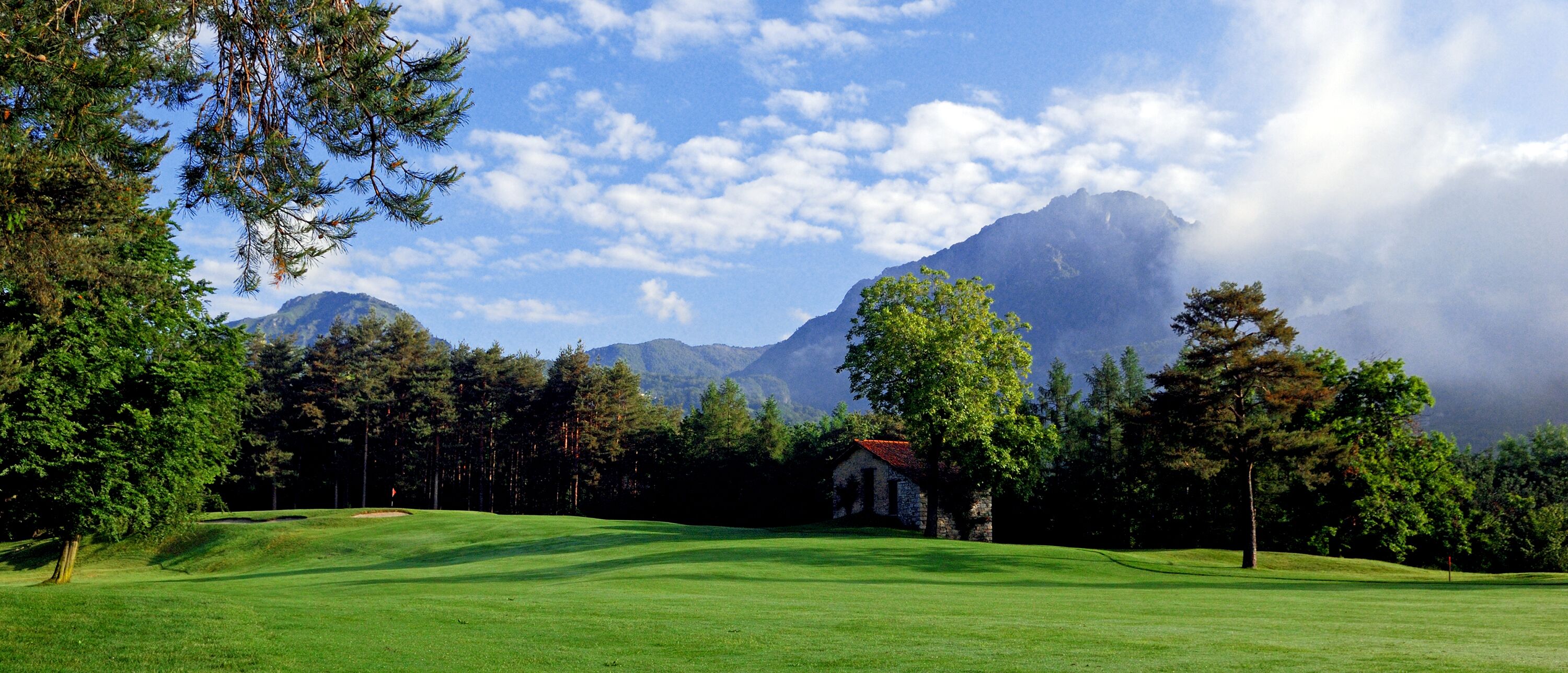 A golf course with a stone building, surrounded by trees and mountains in the background.