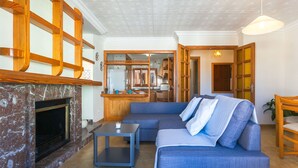 Un salon avec une cheminée, un canapé, une table basse et un coin cuisine visible à travers une porte ouverte.