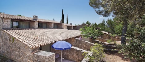A stone building with a tiled roof, a patio with a blue umbrella, and a view of trees and a clear sky.