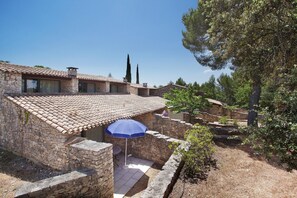 A stone building with a tiled roof, a patio with a blue umbrella, and a view of trees and a clear sky.