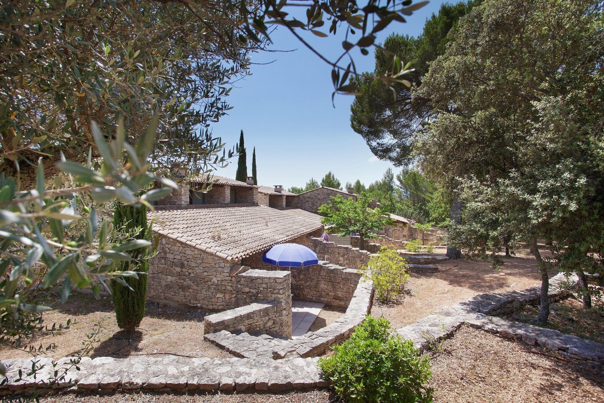 A stone building with a tiled roof, surrounded by trees and shrubs.