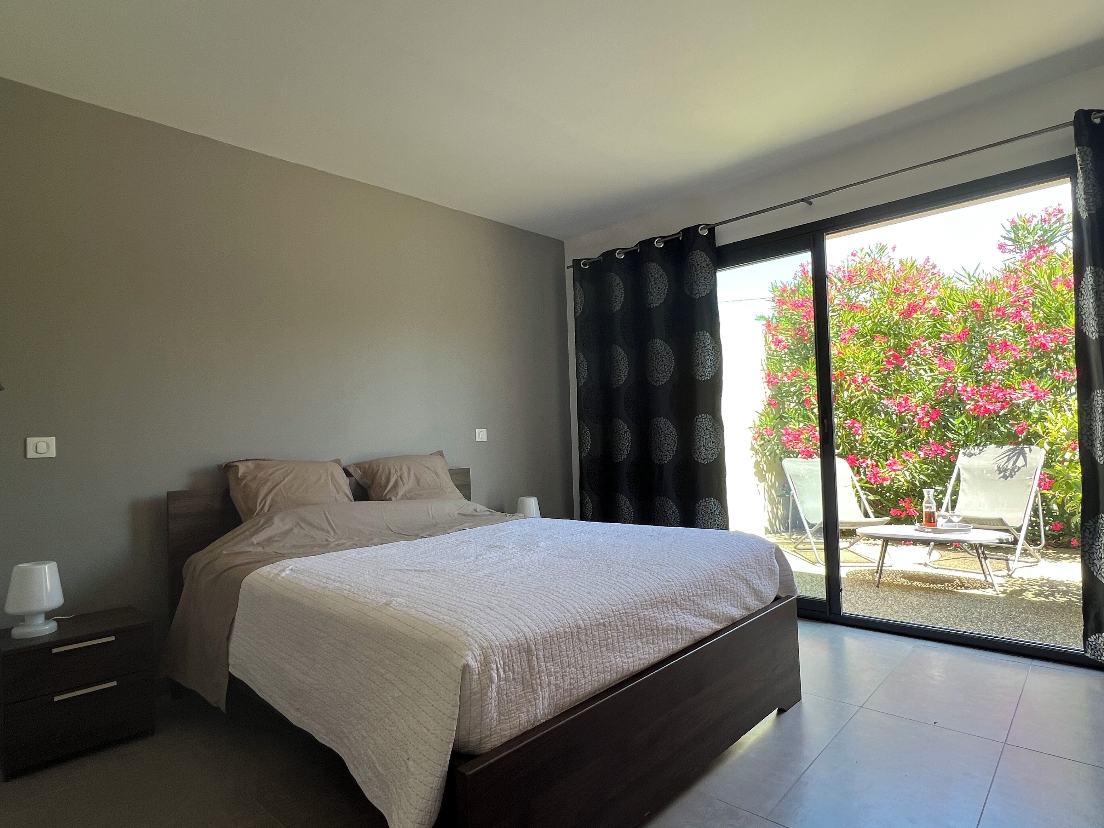 A bedroom with a bed, a nightstand, a lamp, and a view of a balcony with flowers.