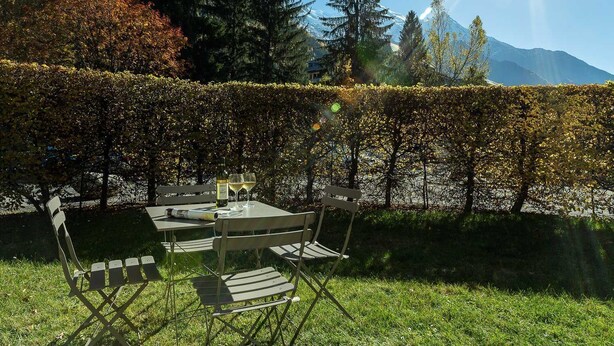 Ein Tisch draußen mit Stühlen, einer Flasche und Gläsern, umgeben von einer Hecke und Bäumen.