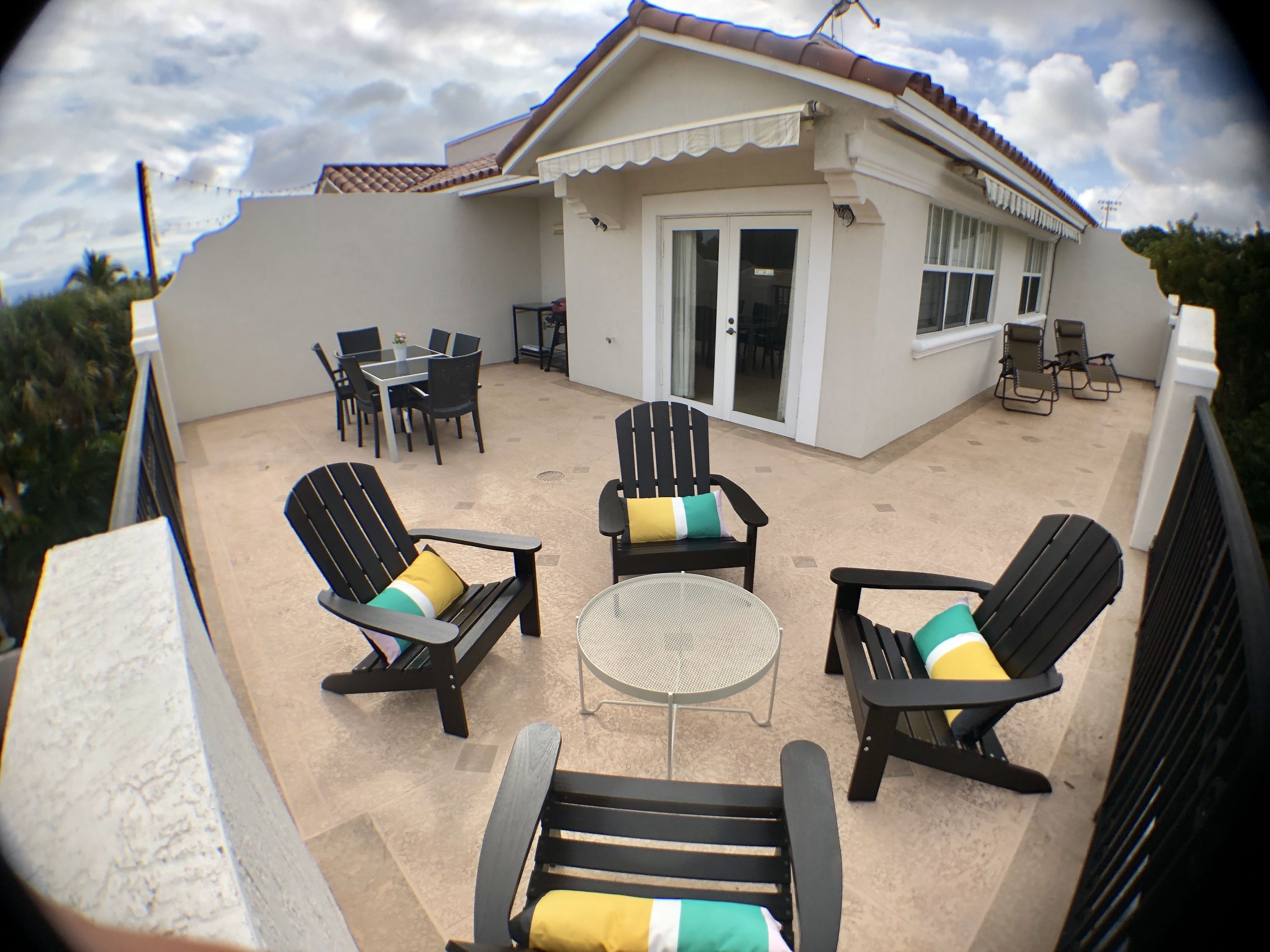 A covered outdoor seating area with chairs and a table, a building with a tiled roof, and a door with glass panels.