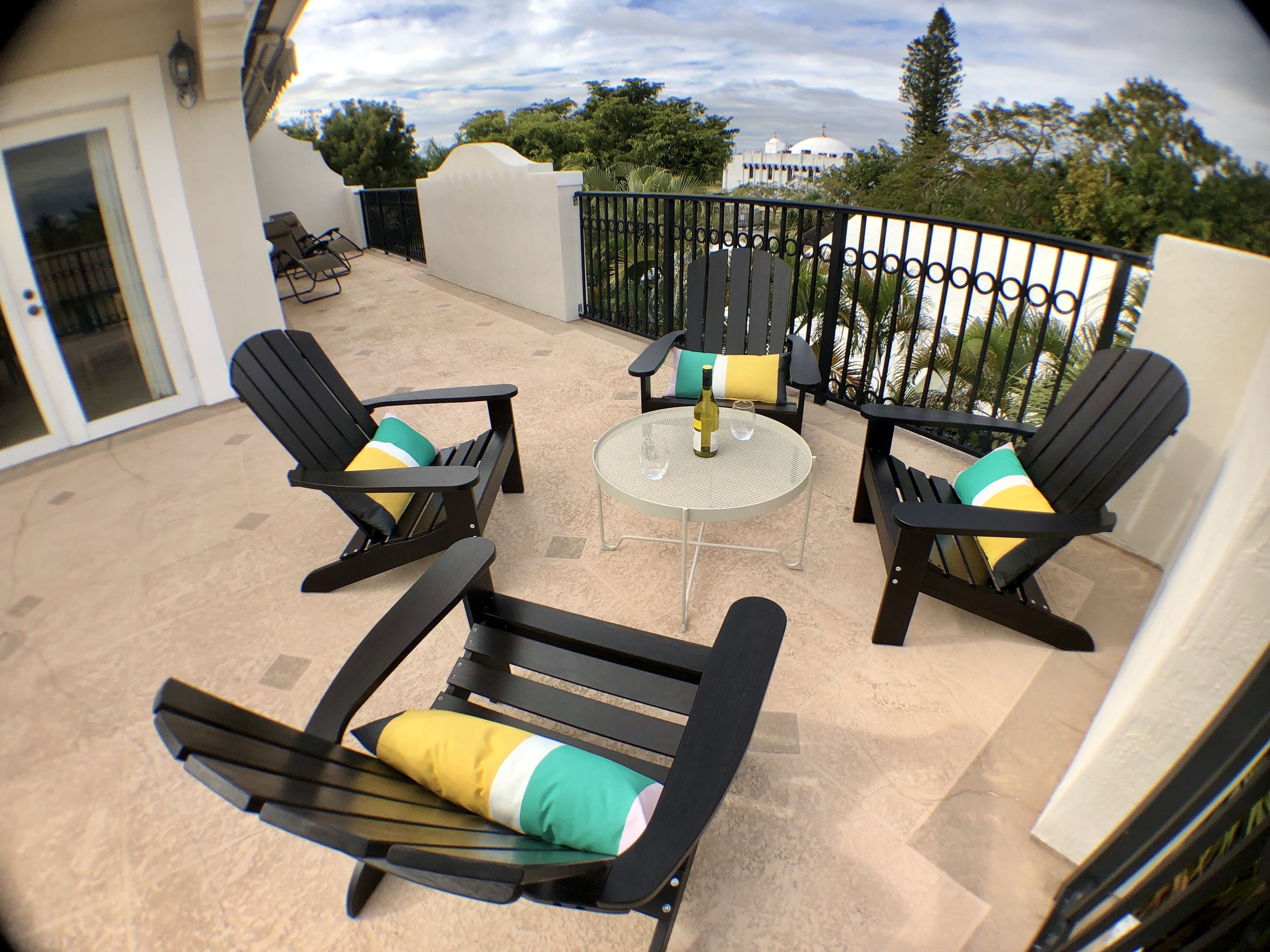 Ein Balkon mit vier schwarzen Liegestühlen, jeder mit einem farbenfrohen Kissen, und einem kleinen runden Tisch mit einer Flasche und Gläsern.