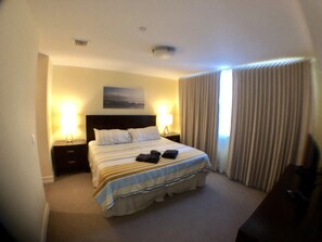 A hotel room with a large bed, two bedside lamps, a headboard, a painting on the wall, and a window with curtains.