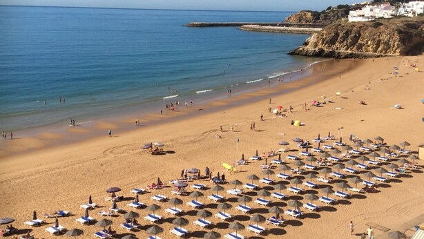 A beach with numerous sun loungers and umbrellas, a clear sky, and a rocky coastline.