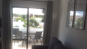 A living room with a dark sofa, a coffee table, and a dining area with chairs and a table. There is a sliding glass door leading to an outdoor patio with a view of buildings and greenery.