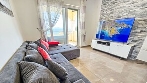 A modern living room with a grey sectional sofa, a flat-screen TV mounted on a white cabinet, and a large window with a view of the sea.