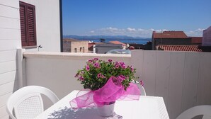 Een balkon met een witte tafel en stoelen, een bloempot met roze bloemen, en een uitzicht op zee en bergen.