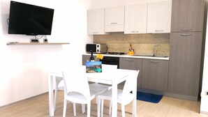 Een compacte keuken met witte kastjes, een magnetron, een fornuis en een spoelbak. Er is een eettafel met stoelen en een flatscreen televisie aan de muur bevestigd.