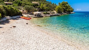 A pebble beach with a row of canoes, a stone wall, and a building with a roof overlooking the water. - Sunny side Beach house (Vela Luka)