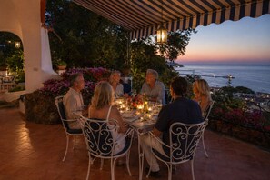 A group of people dining at an outdoor table with a view of the sea and a sunset.