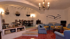 Amplia sala de estar con techo alto, candelabro, zona de comedor con muebles de madera y televisor.