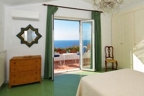 A bedroom with a large bed, a dresser, a chair, and a view of the sea through a sliding glass door.