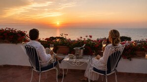 Dos personas sentadas a una mesa con una botella de champán, contemplando la puesta de sol sobre el mar.