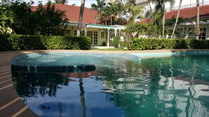 Piscina com borda azulejada, rodeada por vegetação exuberante e um edifício com telhado de telhas vermelhas.