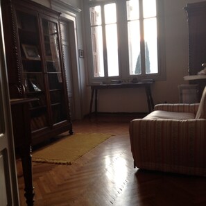 A room with a wooden floor, a patterned armchair, a wooden bookcase, and a window with curtains.
