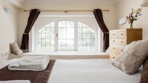 Une chambre avec une grande fenêtre en arc de cercle, un lit recouvert de draps blancs, une commode en bois et un miroir posé sur la commode.