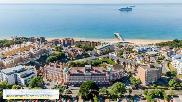 Vue aérienne d’une ville côtière, avec un bâtiment remarquable surmonté d’un dôme vert, une jetée s’avançant dans la mer et un ciel bleu dégagé.