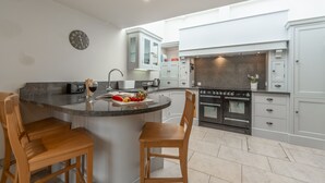 A modern kitchen with a central island, wooden chairs, a clock on the wall, and stainless steel appliances. - Palgrave Barn, Burnham Market, Norfolk (Burnham Market)
