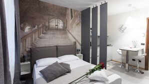 Une chambre à coucher comprenant un lit, une table de chevet, une commode, une lampe fixée au mur, une salle de bain avec un lavabo et un miroir, et un escalier menant à l’étage supérieur.