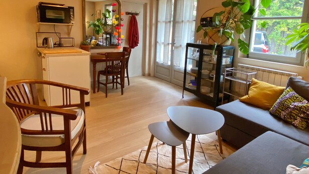 A cozy living room with a sofa, chairs, a small table, and a dining area with a table and chairs. There is a kitchen area with a microwave, a kettle, and a potted plant. A large window provides a view of the outside.