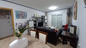 A living room with a sofa, a coffee table, and a TV stand. There is a window with blinds, a painting on the wall, and a vase with flowers on the coffee table. - SPACIOUS APARTMENT WITH WIFI—large terrace and flower-filled garden, sea and mountain views (Adeje)