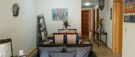 A living room with a sofa, a dining table with chairs, a bookshelf, and a painting on the wall.